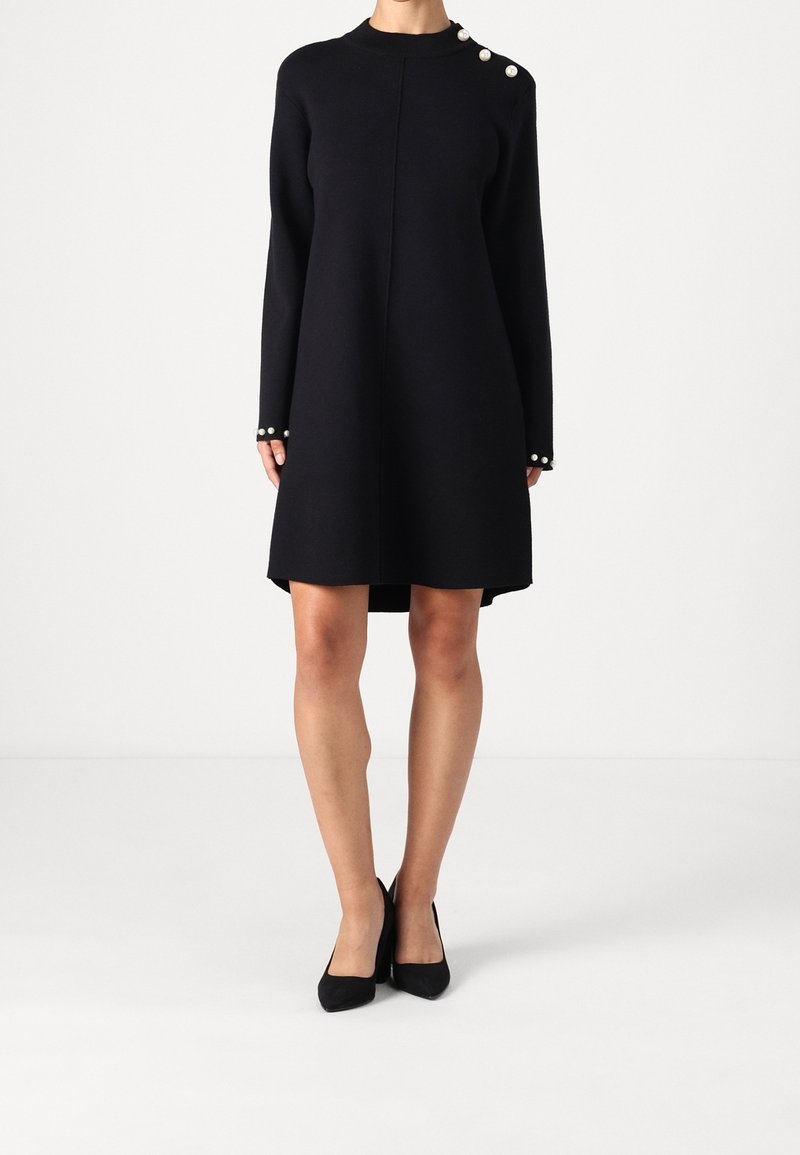 Kvinna i en svart långärmad klänning med pärlknappar på axlarna och vid ärmsluten, tillsammans med svarta klackskor.