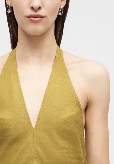 Γυναίκα που φοράει αμάνικη χακί πράσινη μπλούζα με λαιμόκοψη halter και ασημένια σκουλαρίκια κρίκους, με έμφαση στον λαιμό και τους ώμους σε λευκό φόντο.