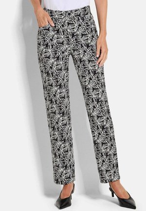 Frau trägt schwarze Hose mit weißem Blumenmuster, kombiniert mit schwarzen spitz zulaufenden Schuhen und einem weißen Oberteil, steht mit den Händen nahe den Taschen.