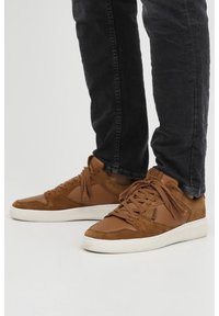 Brown suede and leather sneakers with a white rubber sole, featuring laces, textured accents, and a low-top design.