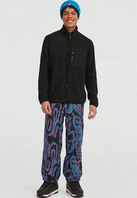 Zwarte zip-up fleecejack met een linker borstzak, gedragen met kleurrijke gestreepte joggingbroeken met blauwe en roze swirlpatronen. Zwarte schoenen.
