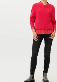 Suéter de punto rosa con textura de canalé, superpuesto a una blusa roja. Combinado con pantalones ajustados negros y botas de tobillo grises con paneles elásticos en los laterales.