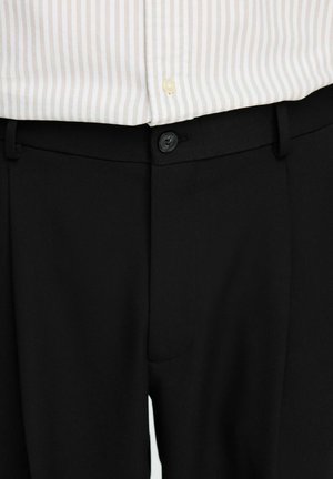 Pantalons noirs ajustés avec une texture lisse, fermeture à bouton et passants de ceinture, associés à une chemise claire à rayures.