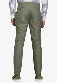 Hombre con pantalones a medida color verde oliva, camisa de rayas y zapatos de ante verdes, visto desde atrás sobre un fondo liso.