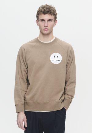 Brown sweatshirt with a round white graphic featuring a neutral expression. Ribbed cuffs and hem; soft, textured material. Simple design.