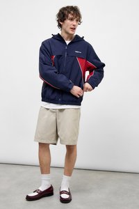 Μπλε σκούρο και κόκκινο αντιανεμικό με κουκούλα, με ελαστικές μανσέτες και τελείωμα. Συνδυασμένο με μπεζ σορτς και σκούρα μοκασίνια με λευκές λεπτομέρειες.