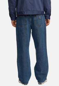 Jean en denim bleu avec une coupe ample, présentant une taille haute, deux poches arrière et un léger délavage. Associé à une veste navy.
