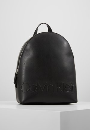 Black leather Calvin Klein backpack with top handle and silver zipper, embossed brand name on front, displayed on a white pedestal.
