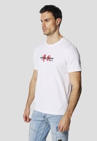 Maglietta bianca in cotone con maniche corte e scollo rotondo, con una stampa grafica rossa e nera della parola "KOROSHAND" sul davanti.