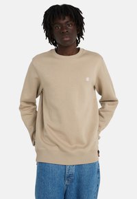 Young person with dark curly hair wearing a beige crewneck sweatshirt and blue jeans, standing against a plain white background.