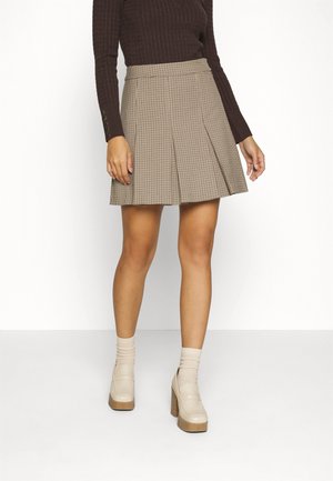 Vrouw draagt een beige en bruin geruite plissérok, donkerbruine top met lange mouwen, beige sokken en beige platformhakken, staand op een witte vloer.