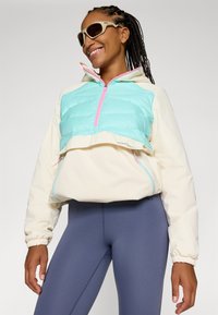 Polstret, kort jakke i mintgrønn og krem med en rosa glidelås, med elastiske mansjetter og sideglidelåser. Kombinert med tettsittende grå leggings.