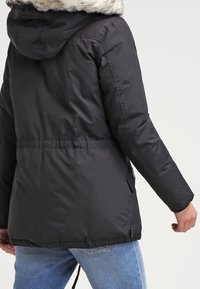 Black puffer jacket with faux fur hood, elastic waistline, long sleeves, side pockets, and textured fabric, worn over blue denim jeans.