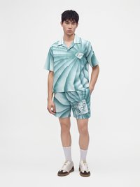 Jeune homme debout portant une chemise à manches courtes à motif spirale bleu sarcelle et blanc ainsi qu’un short assorti avec des patchs à motif visage, des chaussettes blanches et des baskets.