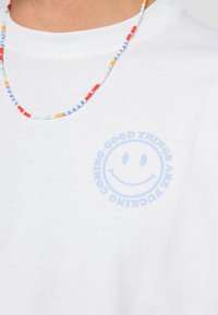 Biely bavlnený tričko s grafikou svetlomodrej usmiatej tváre a textom "DOBRÉ VECI PRICHÁDZAJÚ" v kruhovom dizajne. Viditeľný textúrovaný náhrdelník.