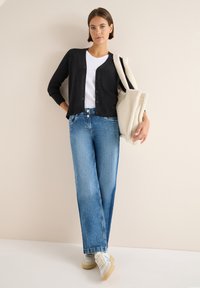 Cardigan nero, t-shirt bianca, jeans dritti blu chiaro e sneakers bianche con suola beige. Porta una borsa a tracolla crema con texture.