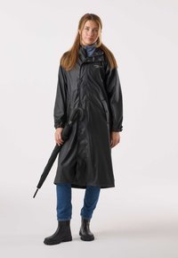 Czarny wodoodporny płaszcz przeciwdeszczowy z zapinaniem na zatrzaski, długimi rękawami i dwoma kieszeniami. Model trzymający czarną parasolkę, ubrany w niebieskie dżinsy i czarne buty.