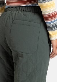 Gequiltte groene broeken hebben een elastische tailleband en een grote achterzak met golvenpatroonstiksel op een gestructureerde stof.
