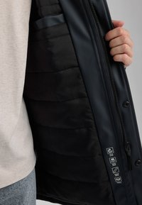 Miestna zateplená bunda so svetlou vonkajšou časťou, čiernou podšívkou a gumovým kovania. Štítky naznačujú vetruvzdorné a vodeodolné vlastnosti.