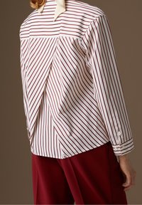 Camisa a rayas en algodón blanco y burdeos, con una abertura en la parte trasera, mangas largas y puños abotonados. Combinada con pantalones burdeos.