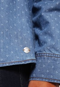 Tessuto chambray blu con piccoli cluster di punti bianchi, caratterizzato da un bottone argento inciso con "SO" e doppia cucitura arancio-marrone su un polsino della manica.