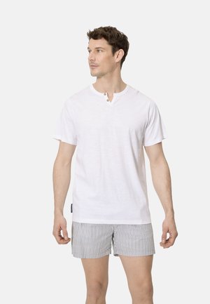 Hombre vistiendo una camisa Henley blanca de manga corta y pantalones cortos a rayas grises y blancas, mirando hacia su izquierda contra un fondo liso.