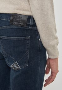 Uomo che indossa jeans Roy Roger's blu scuro e un maglione beige, visibile la tasca posteriore e la patch del marchio sulla vita e sulla tasca.