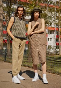 Gilet en tricot pour homme à motif chevron multicolore, joggers beige, et robe sans manches matelassée pour femme en marron avec boutons-pression. Baskets blanches.