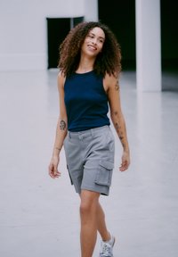 Canottiera blu navy abbinata a pantaloni cargo grigi con tasche laterali, che cammina su un pavimento liscio, con capelli ricci e scarpe da ginnastica.