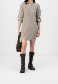 Femme portant une robe beige en tricot à manches longues, des bottes noires montantes et tenant un sac à main vert foncé sur un fond blanc uni.