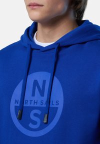 Felpa blu con cappuccio e cordini, caratterizzata da una grande grafica circolare che include il testo "NORTH SAILS" e le iniziali "N" e "S".