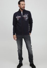 Giacca a zip blu navy con grafiche testuali bianche e rosse, maniche lunghe, colletto rialzato, indossata sopra una maglietta bianca, abbinata a jeans grigi e stivali neri.