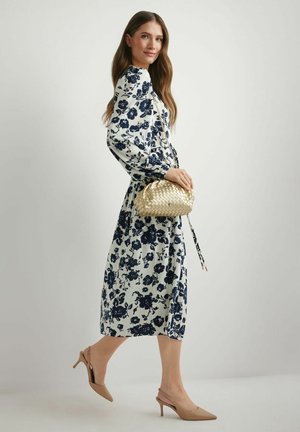 Kobieta w białej sukience midi z granatowym kwiatowym wzorem, niosąca złotą plecioną torebkę, ubrana w beżowe sandały na obcasie z paskiem z tyłu, idąca bokiem.