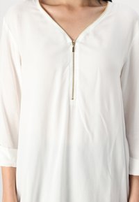 Donna che indossa una blusa bianca con maniche a tre quarti e una zip dorata parzialmente chiusa sul collo.