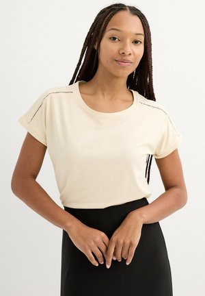 Camiseta de manga corta beige claro con puños enrollados, escote redondo y perforaciones decorativas en los hombros, exhibida con un pantalón negro.