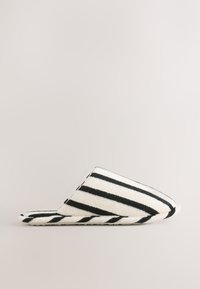 Pantoufle blanche unique avec des bandes diagonales noires, tissu doux et semelle plate, montrée de profil sur un fond neutre.