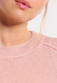 Jasnoróżowy sweter wykonany na drutach, z prążkowanym dekoltem i fakturowanym wykończeniem, z widocznymi detalami szwów na rękawach i dole.