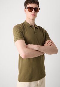 Alyvuogių žalios spalvos polo marškiniai, pagaminti iš medvilnės, su klasikiniu apykaklės kirpimu, trumpomis rankovėmis ir subtiliu logotipu ant krūtinės. Modelis dėvi rudus akinius nuo saulės.