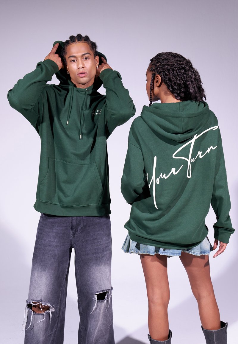 Hoodie vert en tissu doux, avec une poche avant et un grand texte blanc "Votre Tour" au dos. Associé à un jean noir déchiré.
