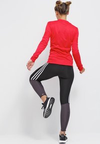 Rød langærmet træningstrøje parret med sorte og grå leggings med hvide sidelister og teksturerede sneakers. Glat, tætsiddende materiale.