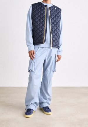 Marineblaue gesteppte Weste über einem hellblauen Sweatshirt, kombiniert mit hellblauen Cargohosen. Blaue Schuhe mit gelben Sohlen. Minimale Design-Details.