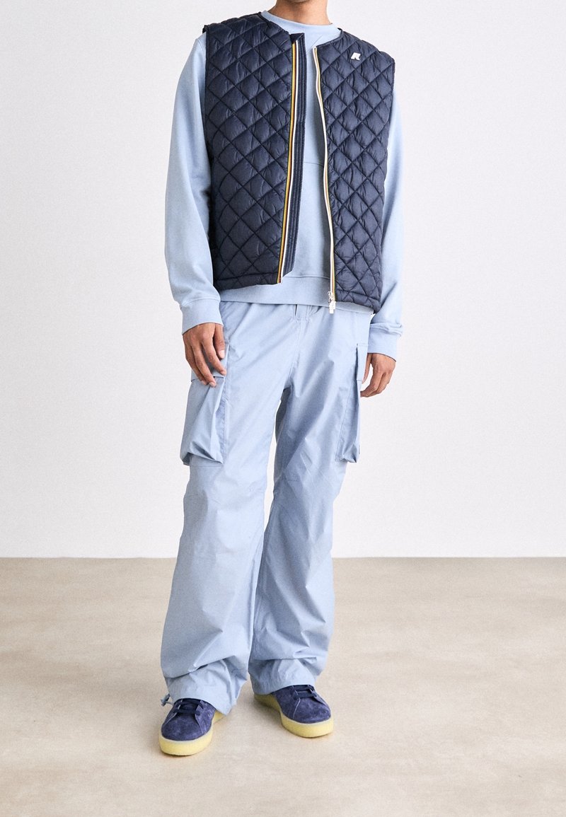 Marineblaue gesteppte Weste über einem hellblauen Sweatshirt, kombiniert mit hellblauen Cargohosen. Blaue Schuhe mit gelben Sohlen. Minimale Design-Details.