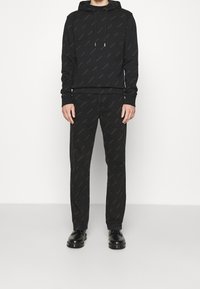 Sudadera negra con un patrón de logo sutil y cordones, combinada con pantalones negros que presentan un patrón similar, complementada con calzado negro.