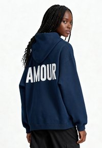 Tumši zila kapuce no kokvilnas maisījuma ar baltu uzrakstu "AMOUR" aizmugurē, ar kapuci un ribotām aprocēm, brīva piegriezuma.