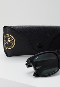 Ochelari de soare Ray-Ban negri cu lentile întunecate, cadru pătrat și detaliu cu logo auriu. Însoțiți de o carcasă texturată neagră cu un emblemă aurie.