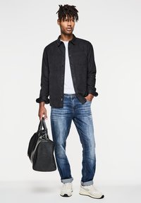 Svart knapparskjorta med bröstficka, vit t-shirt under, blå jeans, svart duffelväska och ljusa sneakers.