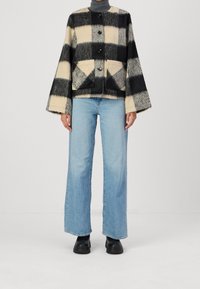 Czarna i kremowa, w kratę, wełniana kurtka z oversizedowymi rękawami i czterema czarnymi guzikami, zestawiona z jasnoniebieskimi szerokimi dżinsami i czarnym obuwiem.