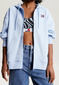Mujer vistiendo una sudadera con cremallera de color azul claro sobre un sujetador deportivo de estampado en blanco y negro, combinada con unos jeans de tiro alto azul.