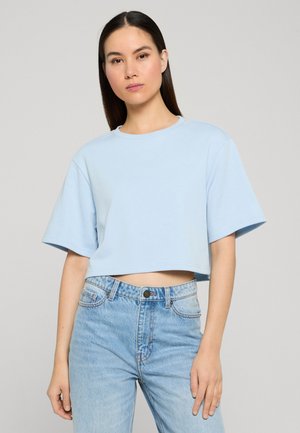 Młoda kobieta z długimi ciemnymi włosami ubrana w jasnoniebieski krótki T-shirt i jasnoniebieskie dżinsy z wysokim stanem na prostym tle.