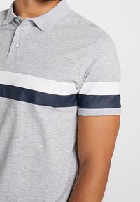 Polo gris avec un col, présentant des rayures horizontales bleu marine et blanches sur la poitrine. Fabriqué en un tissu lisse et texturé.